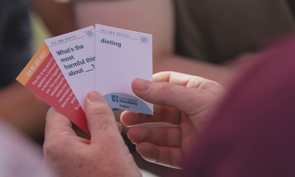A white hand holds three cards from a card game called Conversations Against Mundanity