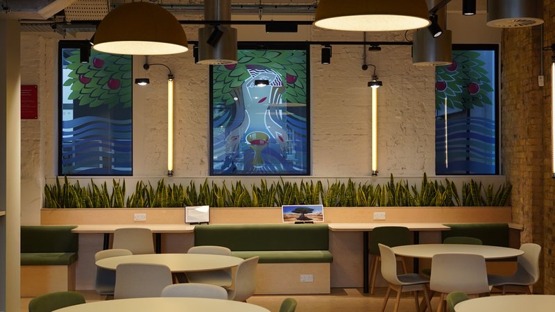 Café‑style seating with round tables, green benches, hanging lamps and colourful communion‑themed murals in the windows.