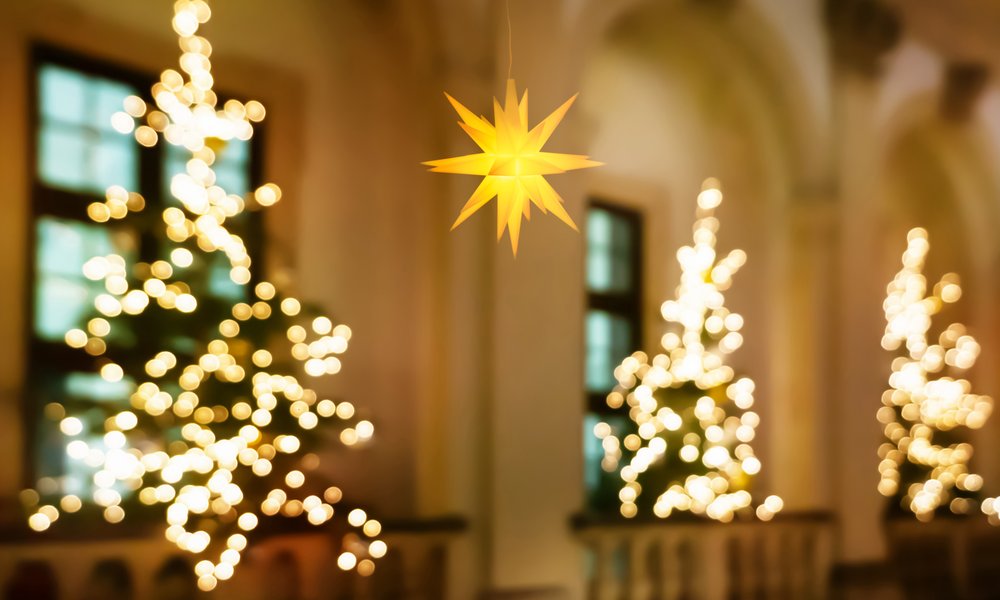 Three Christmas trees inside of a church, in front of windows, with a light hung in the foreground.