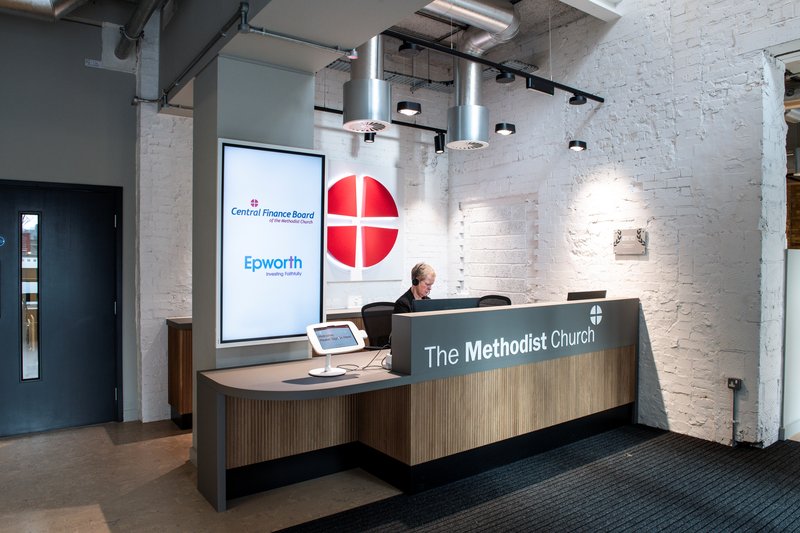 Reception desk at Methodist Church House with staff member seated; large Methodist Church logo and finance‑board display behind.