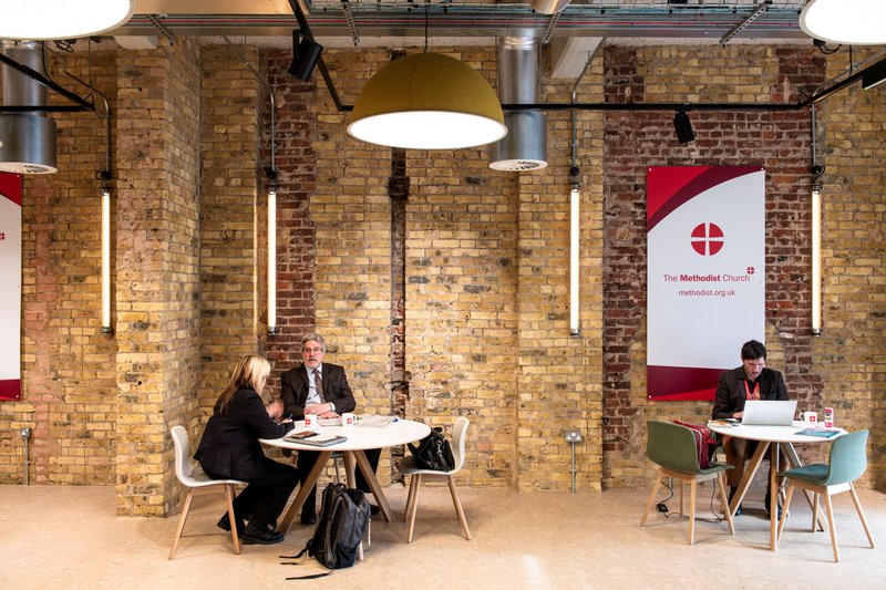 Brick‑walled breakout area where two people meet at one round table and another works on a laptop at a second table.