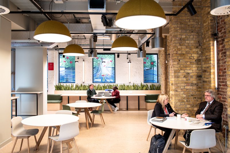Wide view of the communal workspace with round tables, green hanging lamps and colleagues chatting or working.