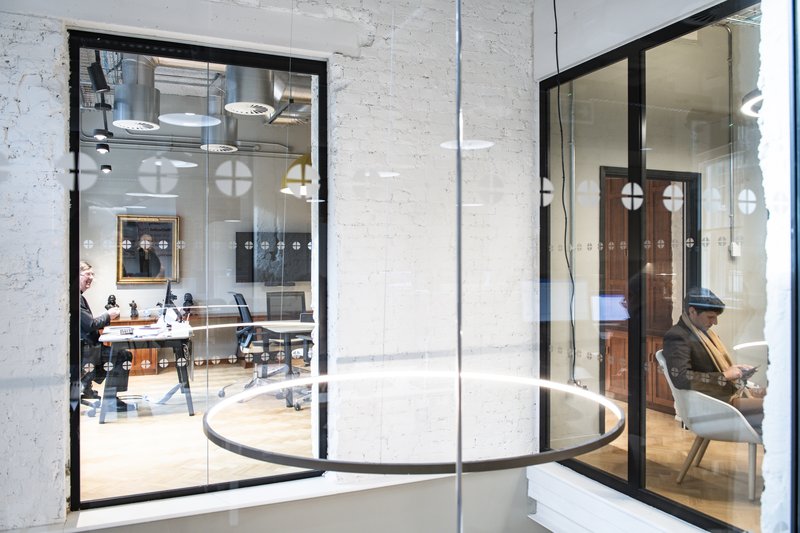 View through glass partitions into two adjacent rooms at Methodist Church House: one person sits at a desk with small bust sculptures, while another sits in a lounge chair using a phone, set against white‑brick walls and exposed ducting.