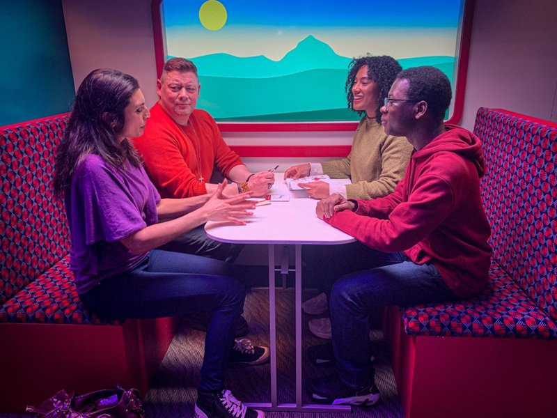 A group of four people on a train journey. They are sitting at a table, having a conversation.