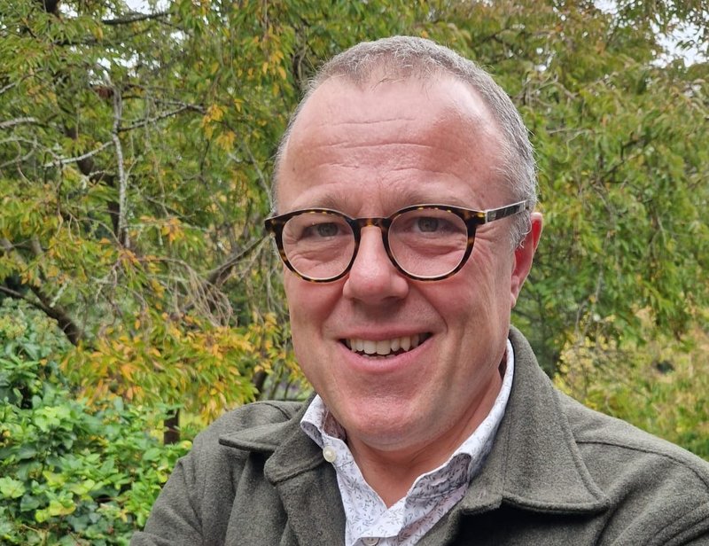 A man wearing glasses standing in front of a bush