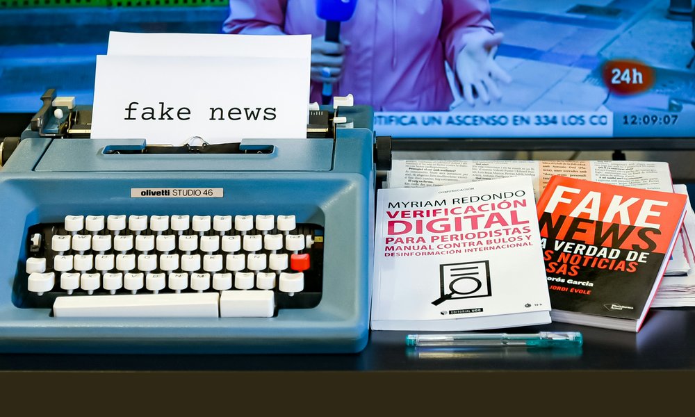 A sheet of paper in a typewriter says 'fake news'. Books on the table also refer to fake news