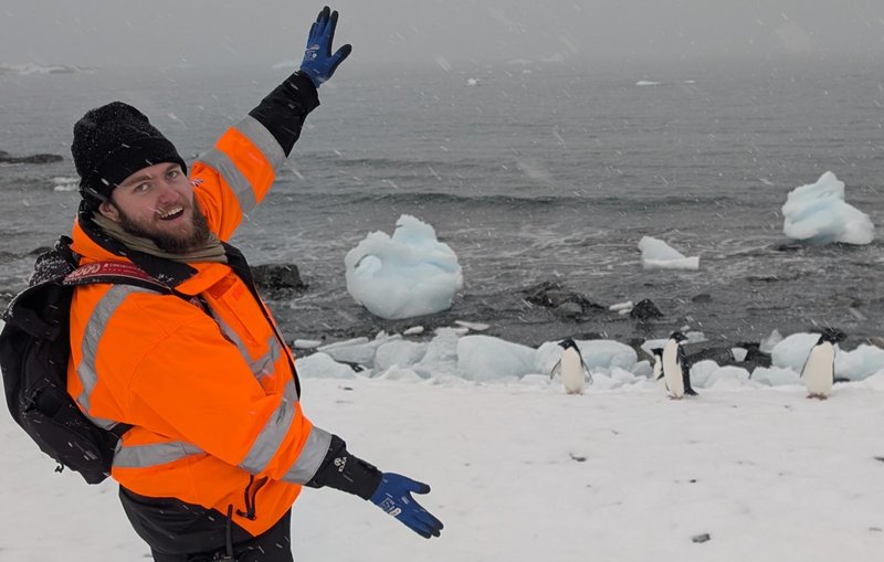 A man dressed for the snow stands at water's edge with penguins