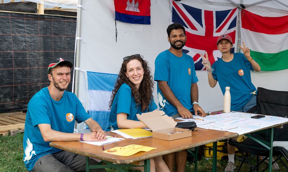four young adults in blue t-shirts sat at a table. Flags of the world behind them