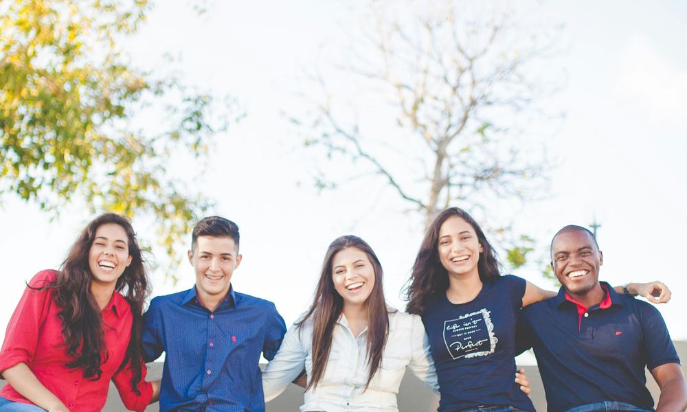 five young adults sit on a wall shelf outdoors, arms round each other, smiling at the camera